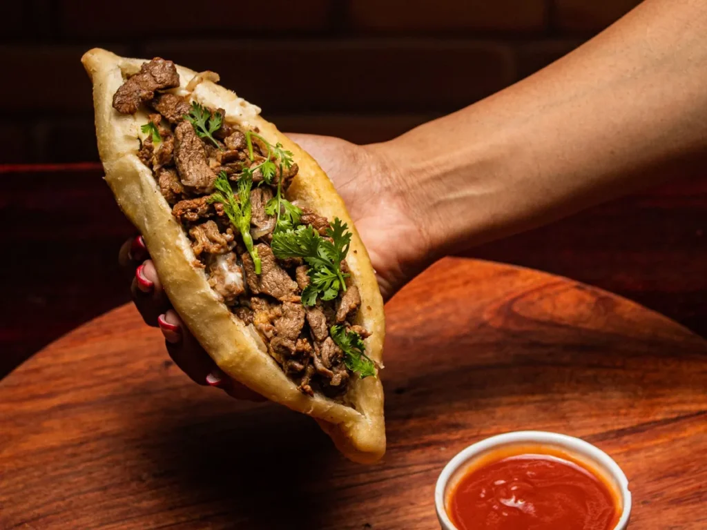 A hand holding a flatbread pocket filled with grilled meat and fresh herbs over a wooden table.