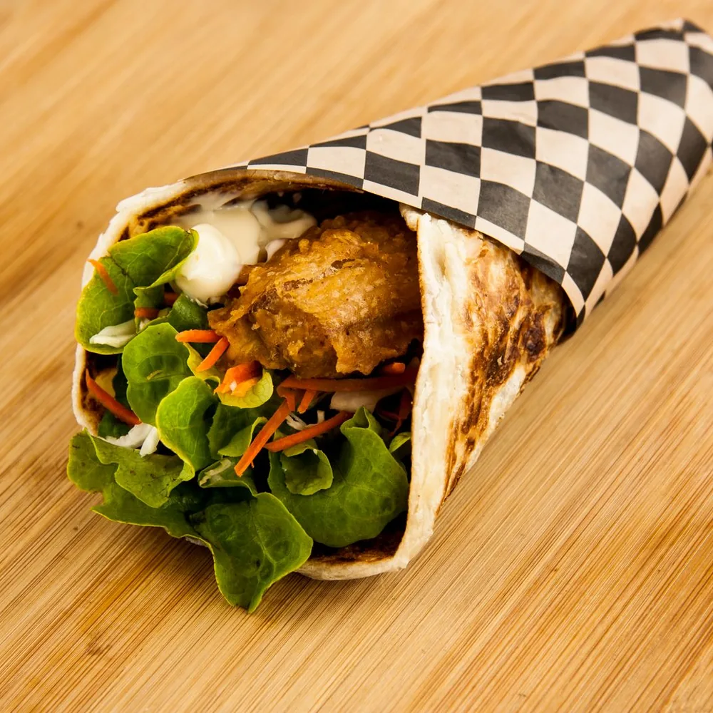 A wrapped flatbread filled with fried chicken, lettuce, and shredded carrots on a wooden board.