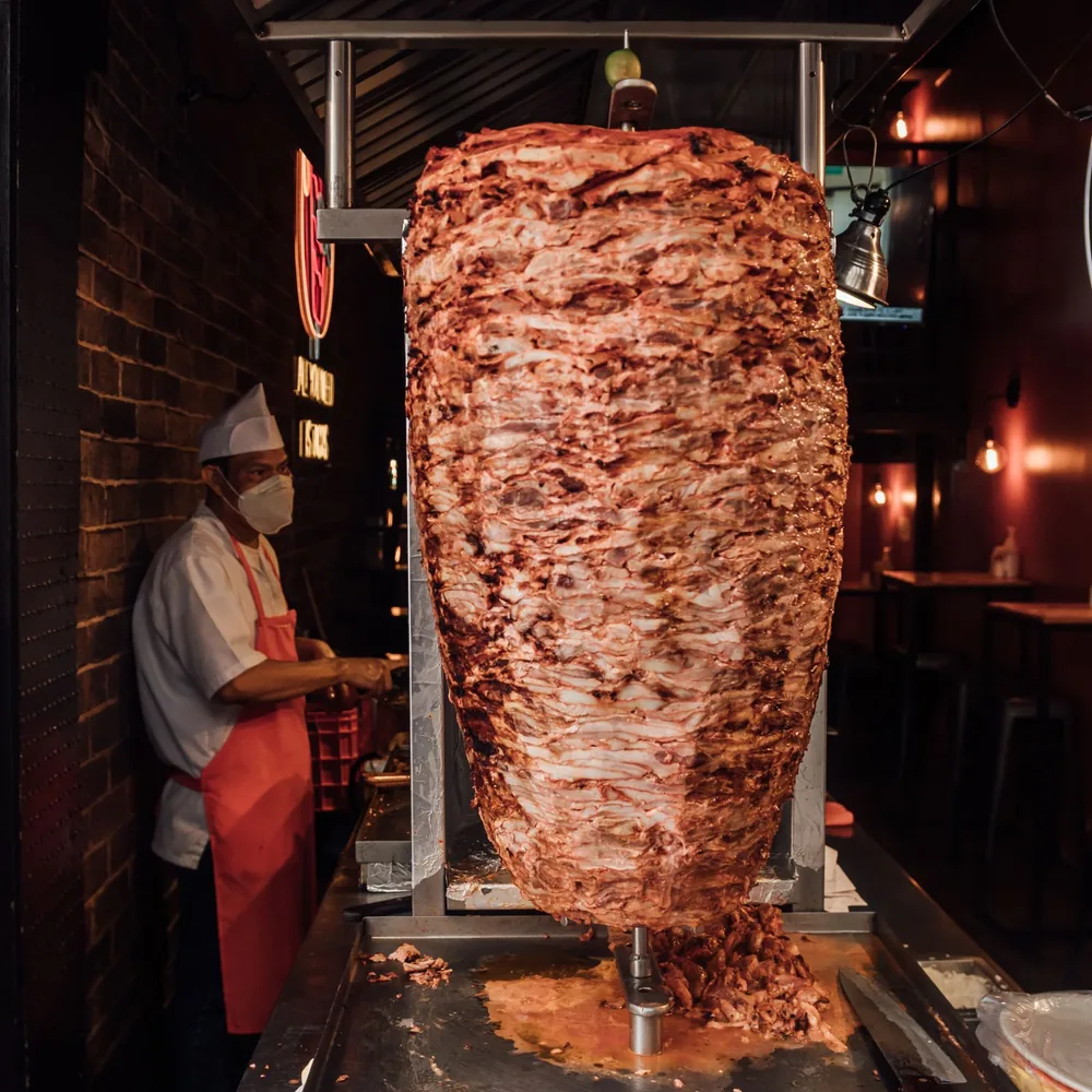 A tall vertical spit of roasted meat in a restaurant with a worker standing nearby.