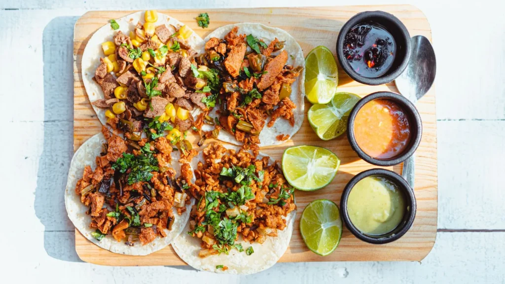 Four tacos on a wooden board with lime halves and three small bowls of sauce.