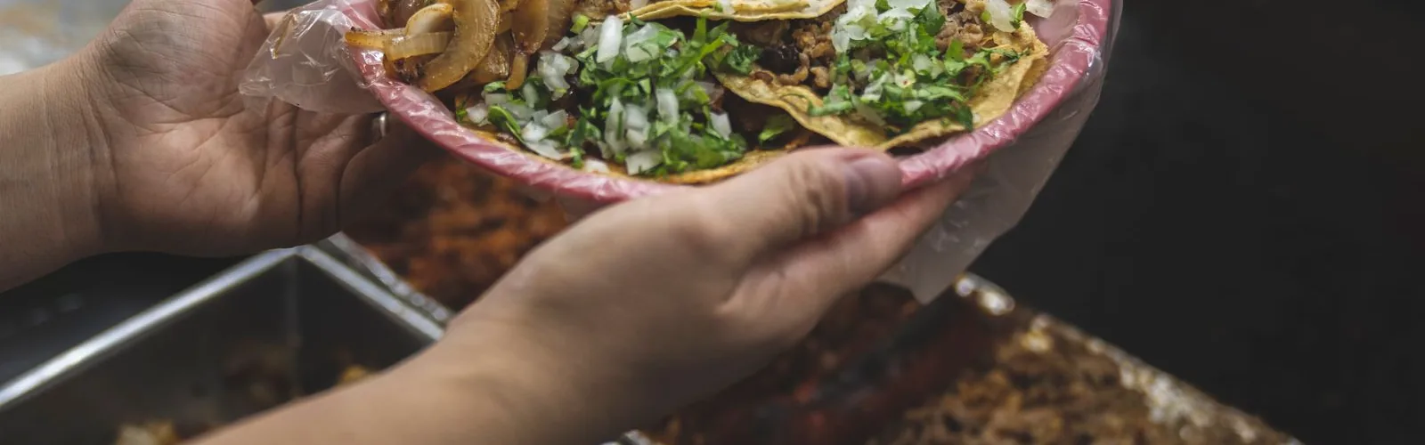 Hands holding a plate of tacos topped with diced onion and cilantro over a serving tray.