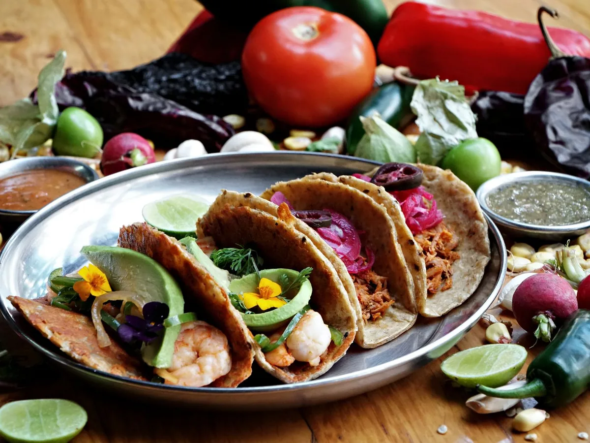 A plate of tacos with shrimp, avocado, and pickled onion surrounded by peppers, tomatoes, and lime wedges.