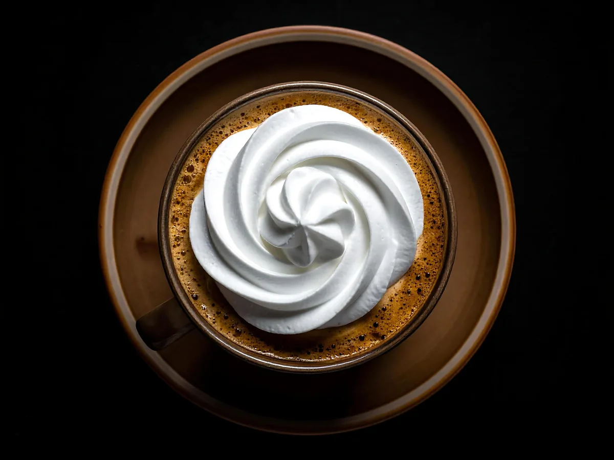 A cup of coffee topped with a swirl of whipped cream, seen from above on a saucer.