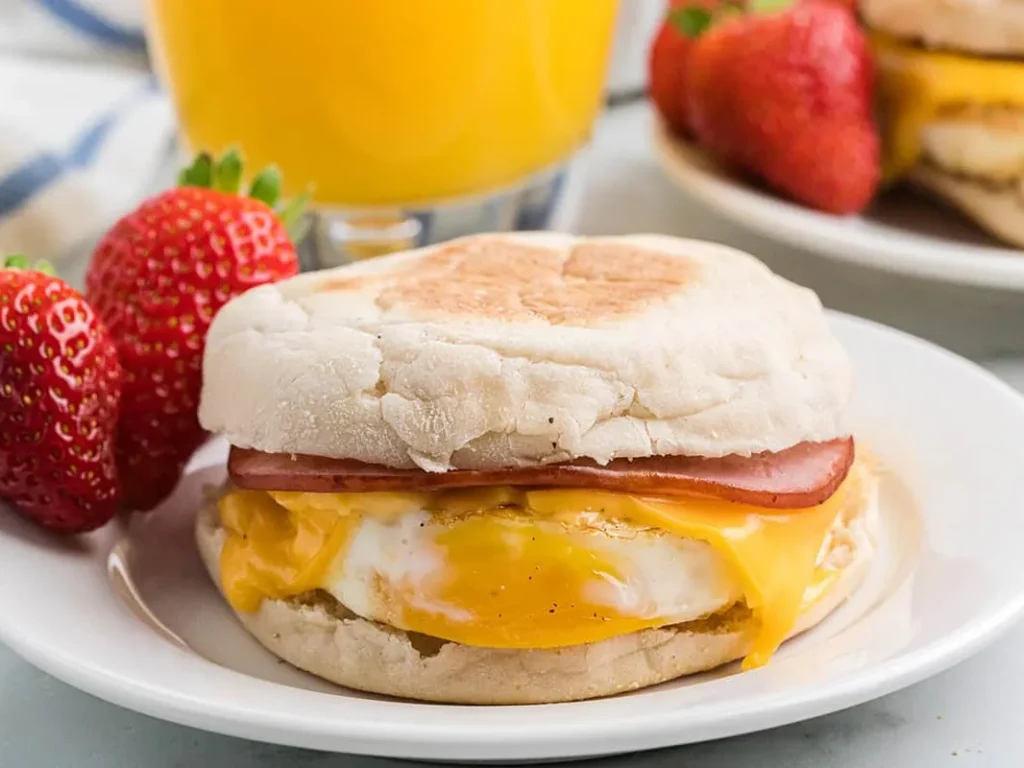 An egg, ham and cheese breakfast sandwich on a muffin beside strawberries, with orange juice behind it.