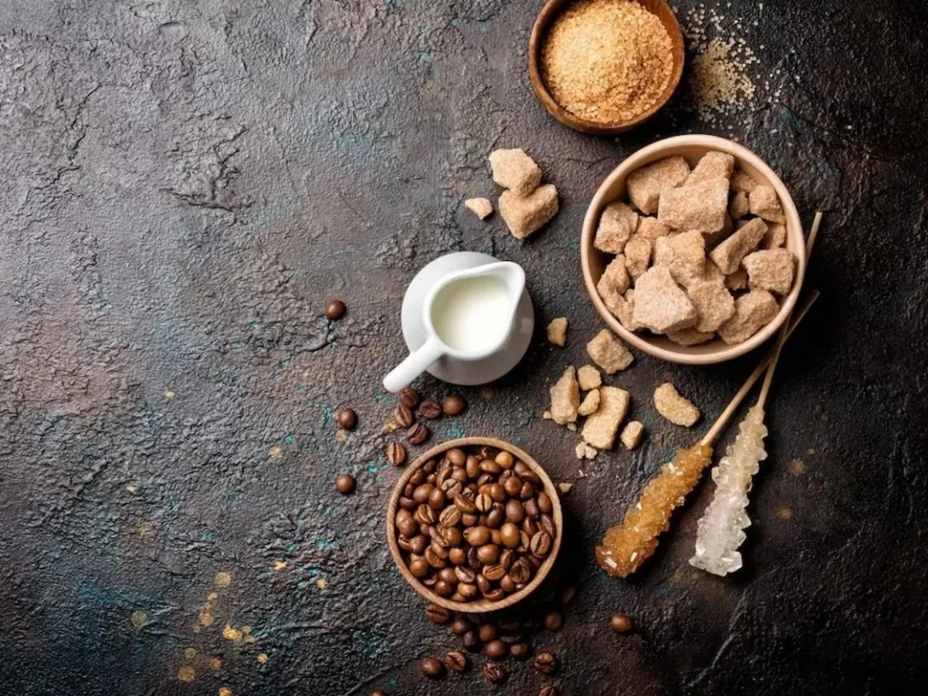 Bowls of brown sugar cubes, coffee beans and granulated sugar beside a small jug of milk on a dark surface.