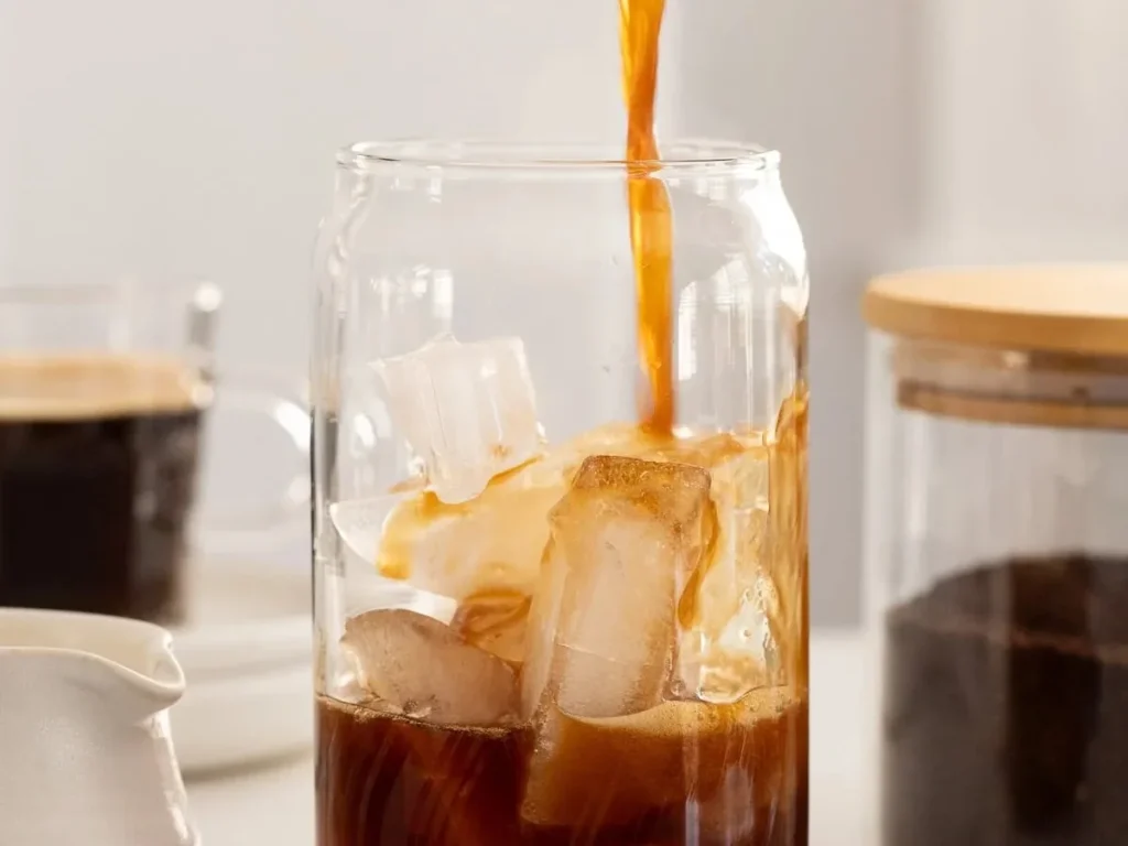 Coffee being poured over ice cubes in a clear glass jar.