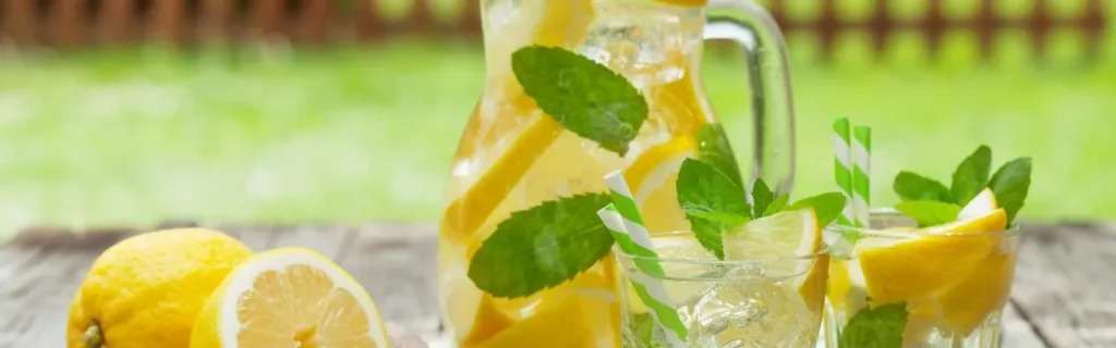 A pitcher and glasses of lemonade with lemon slices and mint leaves on a wooden table outdoors.