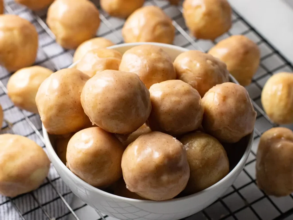 Glazed donut holes piled in a bowl on a cooling rack, with more scattered around it.