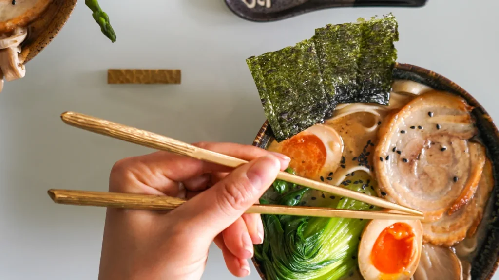 A hand holding chopsticks over a bowl of noodle soup with egg, greens, sliced meat, and seaweed.