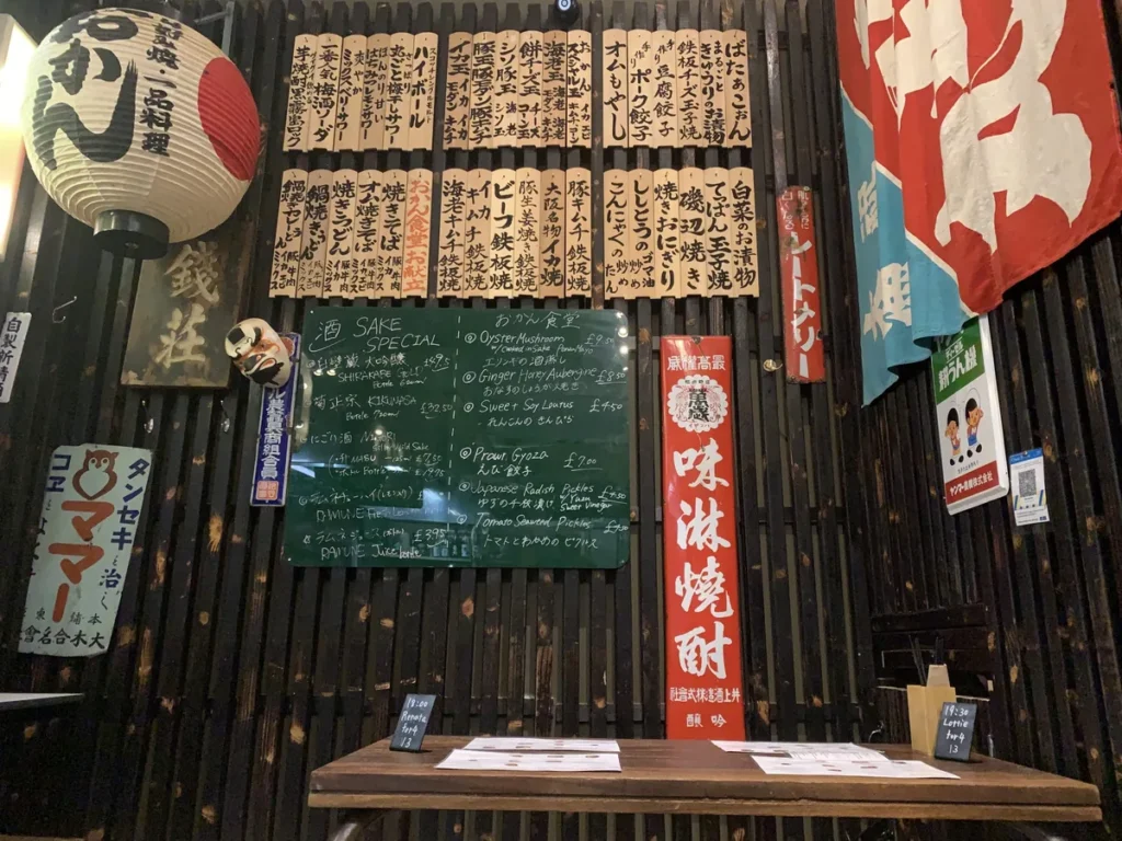 A wooden wall covered with hanging menu strips, a chalkboard menu, banners, and a paper lantern above a table.