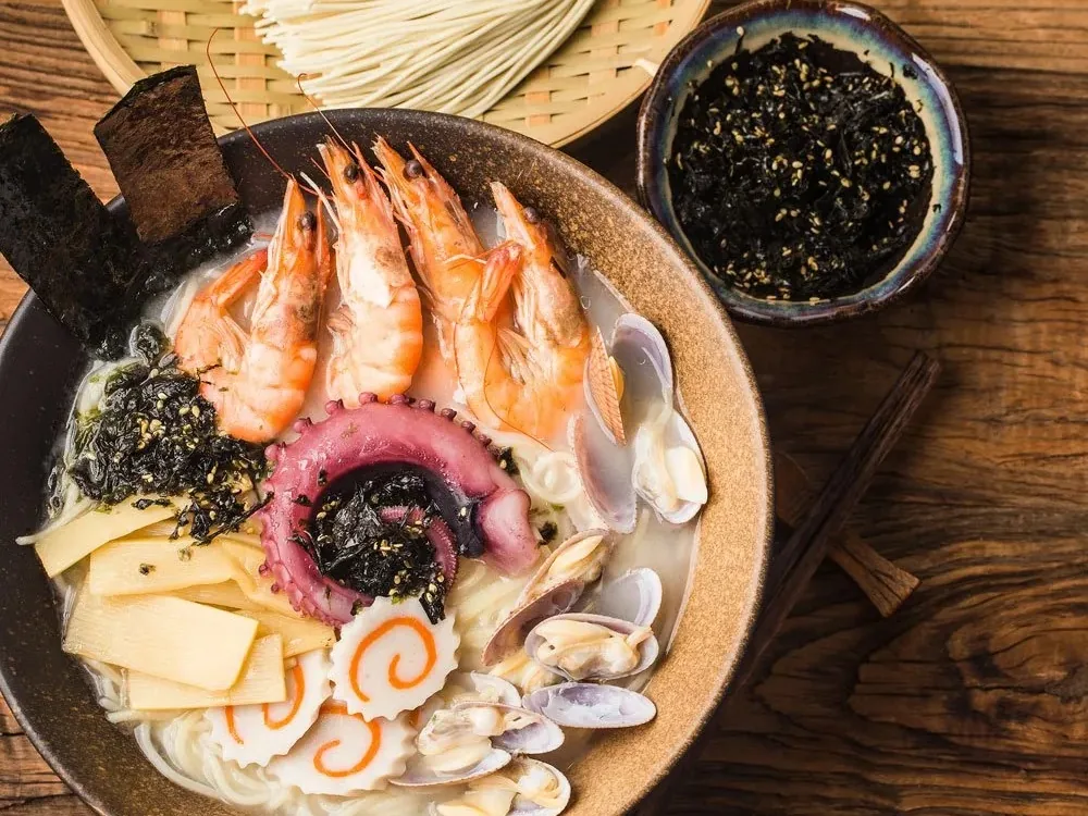 A bowl of seafood noodle soup with prawns, octopus, clams, and seaweed beside a small bowl of seaweed salad.