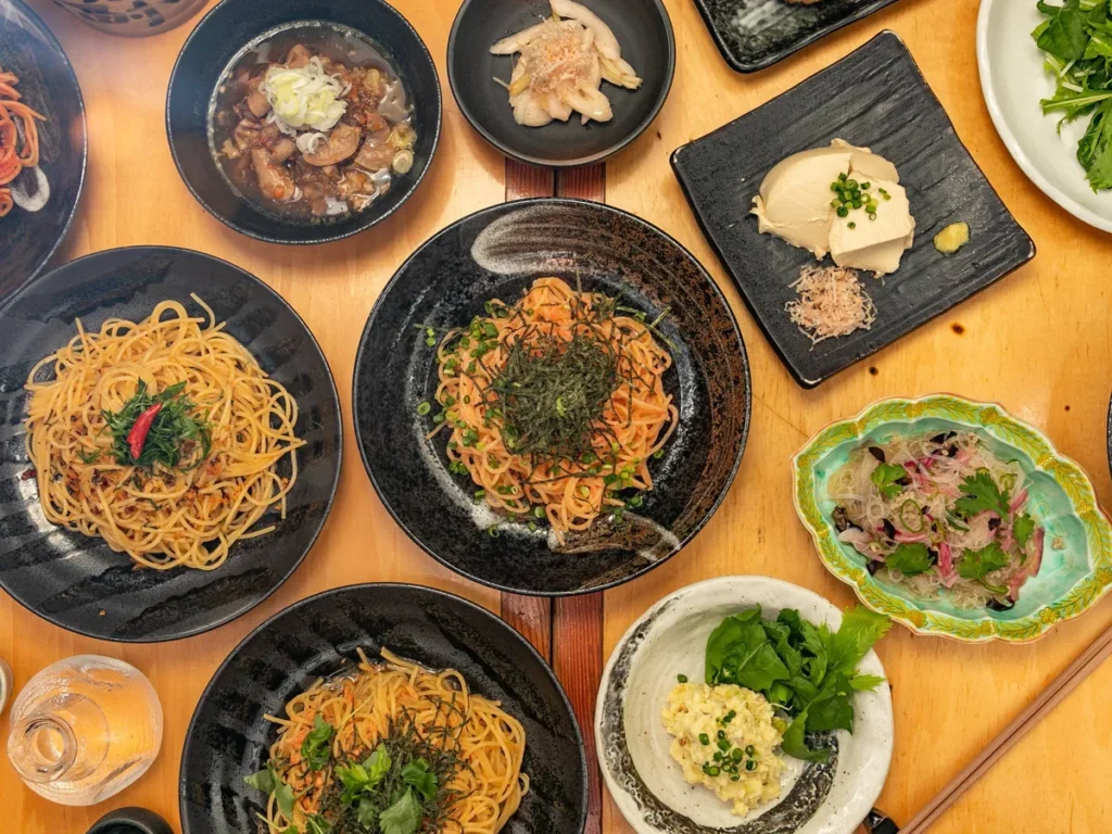 Several bowls of noodle and rice dishes with chopsticks arranged on a wooden table, viewed from above.
