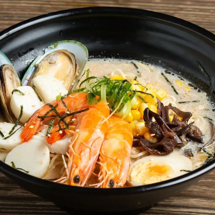 A bowl of seafood noodle soup with prawns, mussels, a halved egg, sweetcorn, and herbs.